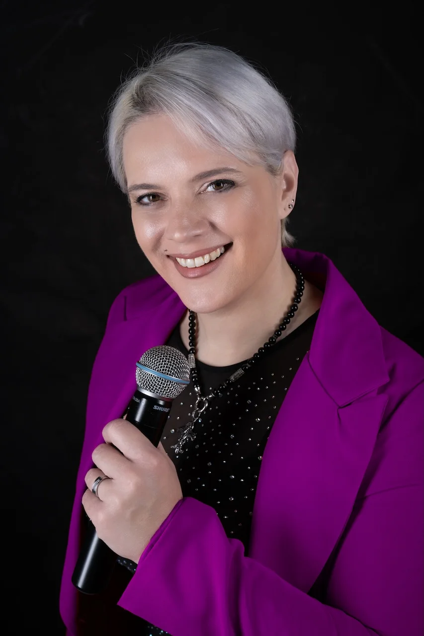 Portrait von Sarah-ANN – Sängerin mit Mikrofon im pinken Blazer, frontal lächelnd