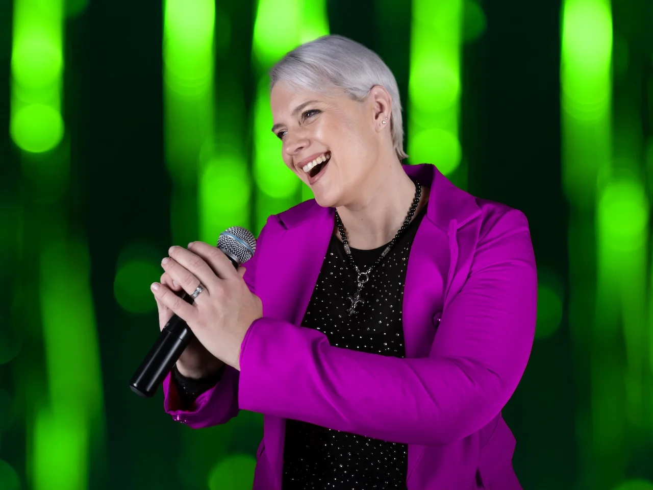 Portrait von Sarah-ANN – Sängerin mit Mikrofon im pinken Blazer, lachend vor grünem Lichterhintergrund