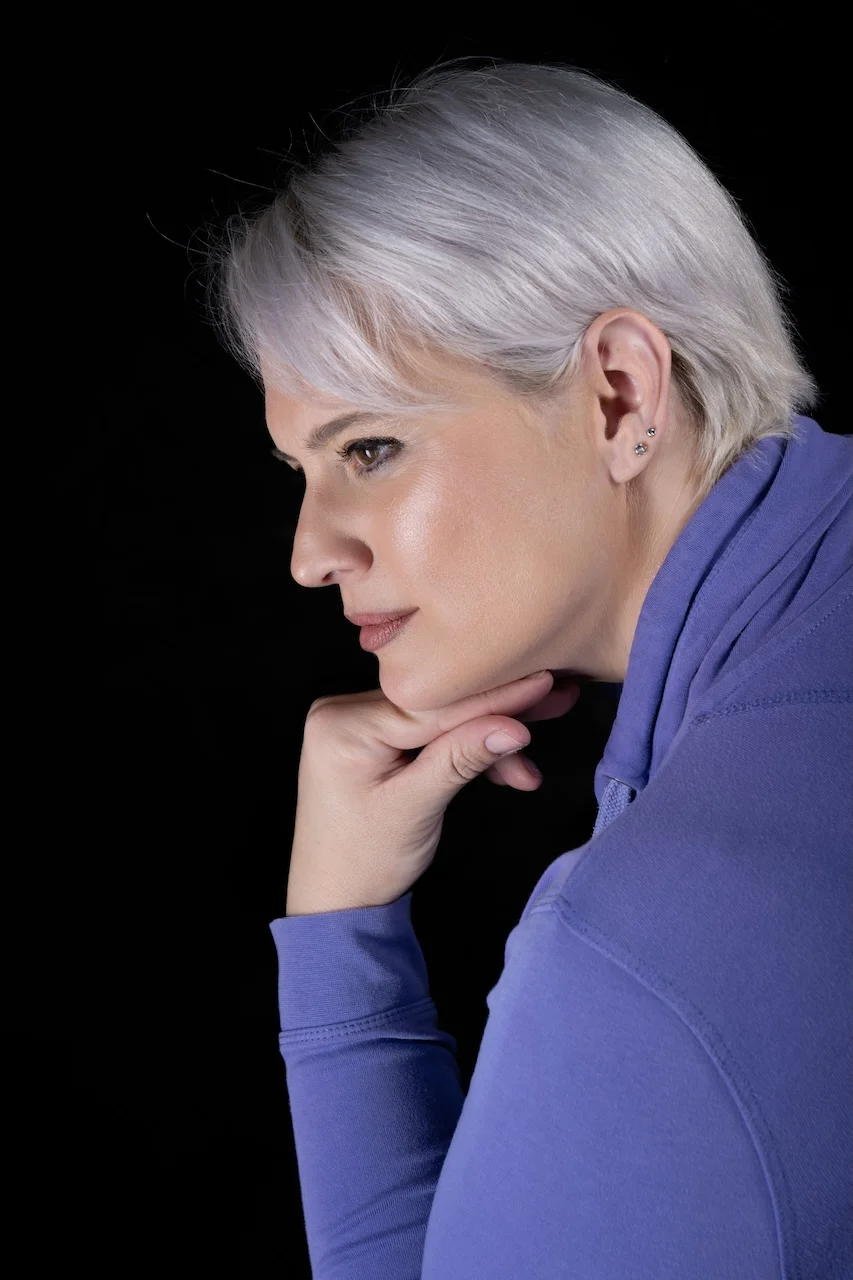 Porträtaufnahme von Sarah-ANN im Profil vor schwarzem Hintergrund, mit kurzen silbergrauen Haaren und nachdenklichem Blick, Hand stützt das Kinn.