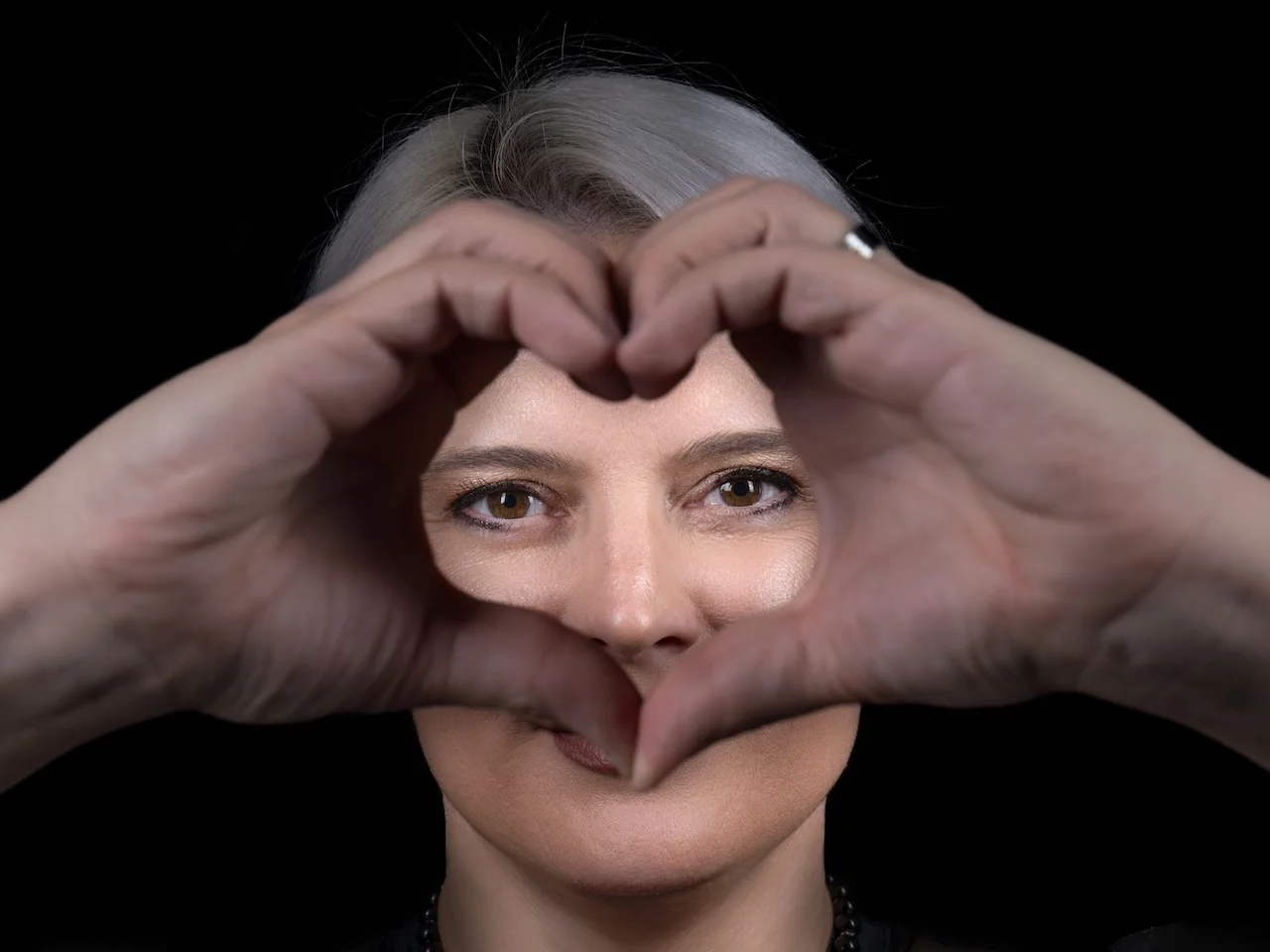 Nahaufnahme von Sarah-ANN, die mit beiden Händen ein Herz formt und durch die Öffnung lächelt, schwarzer Hintergrund.