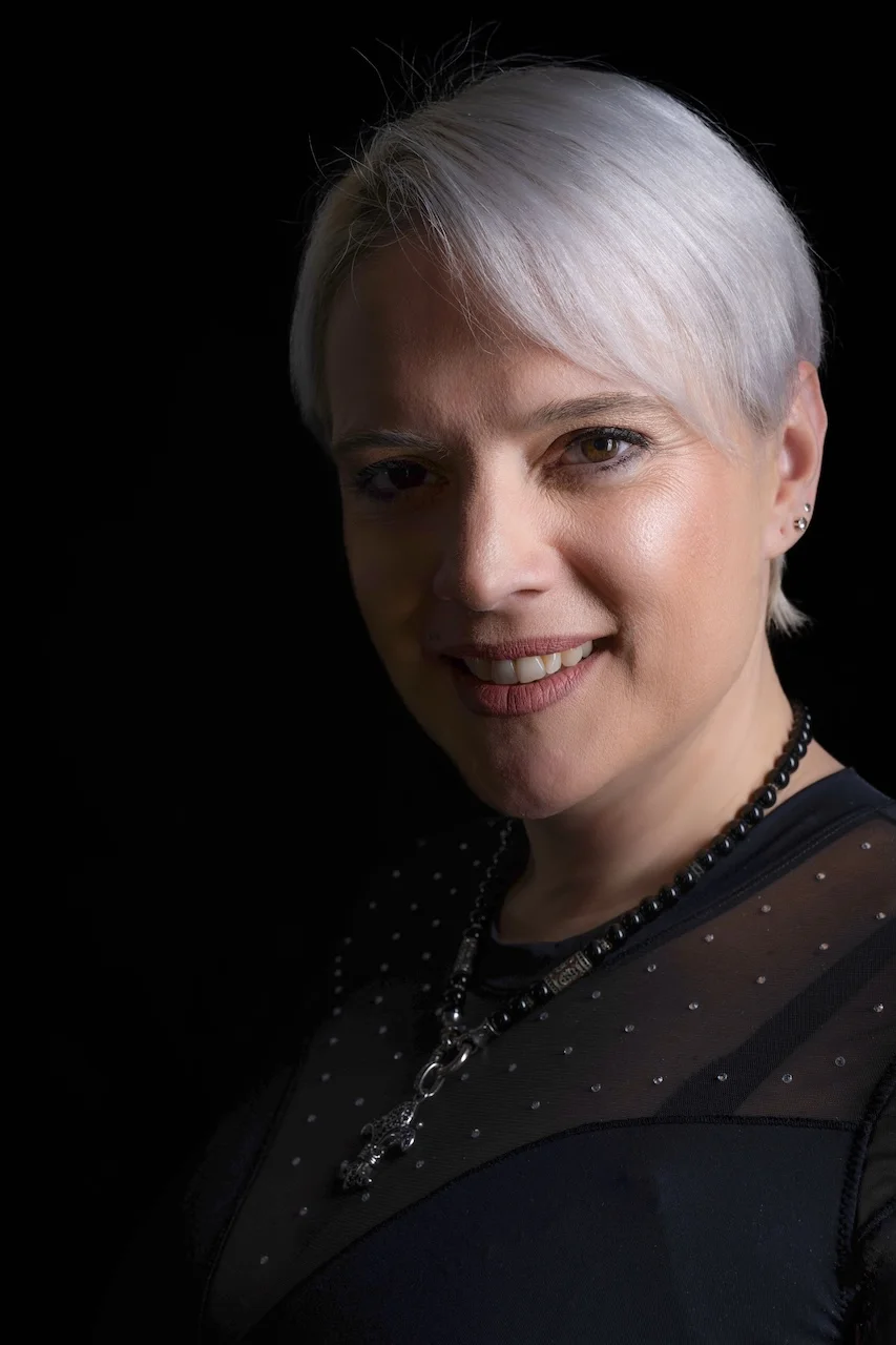 Porträt von Sarah-ANN mit kurzen silbergrauen Haaren, schwarzem Oberteil mit Perlenapplikationen und schwarzer Kette, vor schwarzem Hintergrund, lächelnd in die Kamera.